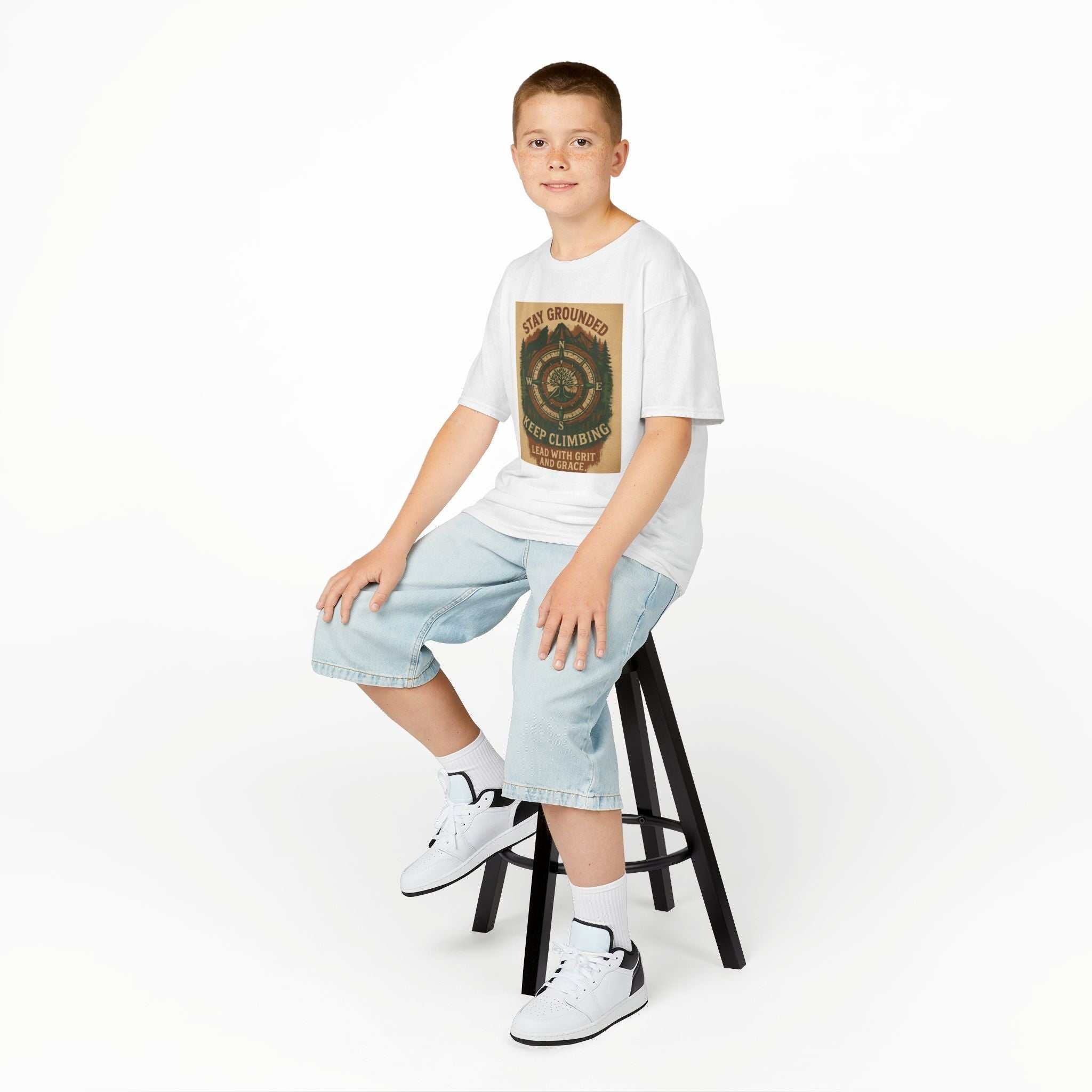 Nature-inspired Adventure Kids Tee with motivational "Stay Grounded & Bring The Good Energy" print, worn by a child seated on a stool.
